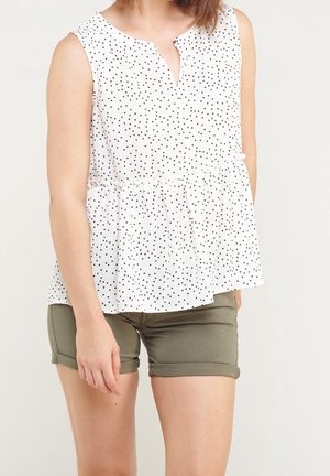 Weißes ärmelloses Oberteil mit schwarzen Punkten, Peplum-Design und einem Schlüssel-Ausschnitt. Kombiniert mit olivgrünen Shorts mit umgeschlagenem Saum.