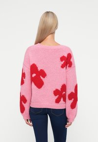 Mulher com cabelo loiro longo, vestindo um suéter rosa de malha com grandes padrões de flores vermelhas, vista de costas, combinado com jeans azul escuro.