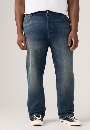 Homme portant un jean bleu délavé et des baskets noires et blanches, debout avec les mains relâchées le long du corps, devant un arrière-plan uni.