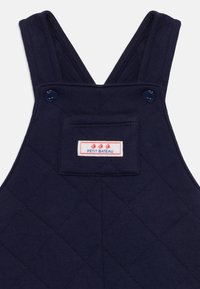 Temno modra prešita kombinezon s naramnicami, ki ima modre gumbe in žep na prsih z rdečo in belo etiketo Petit Bateau.