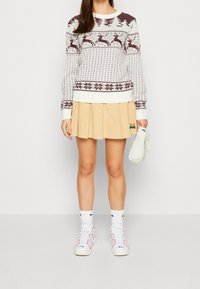Pull blanc tricoté avec des motifs de rennes bordeaux, associé à une jupe plissée beige. Baskets blanches avec des accents rouges.