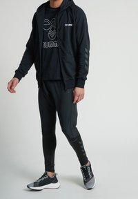Un hoodie noir zippé avec des poignets côtelés, associé à un pantalon noir ajusté. La tenue comporte un t-shirt noir à motif et des chaussures de sport grises.