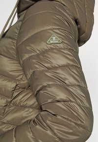 Padded jacket v matné olivově zelené barvě, s prošívanými částmi a trojhrannou nášivkou na rukávu. Měkká textura.