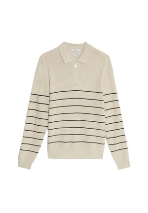 Uomo che indossa camicia beige abbottonata sopra polo beige a righe, pantaloni in velluto a coste neri e scarpe di pelle nere, in piedi con le mani in tasca.