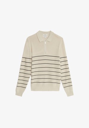 Homme portant une chemise beige boutonnée sur un polo beige rayé, pantalon en velours côtelé noir et chaussures en cuir noires, debout les mains dans les poches.