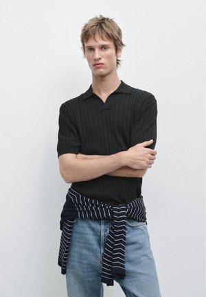 Jonge man met kort lichtbruin haar, gekleed in een zwarte, gebreide korte mouwen shirt en een gestreepte trui om zijn middel geknoopt.