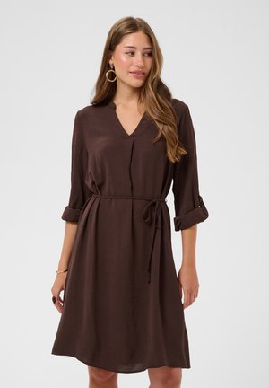 Femme aux longs cheveux portant une robe marron à col en V, manches retroussées et ceinture nouée à la taille, regardant légèrement sur le côté.
