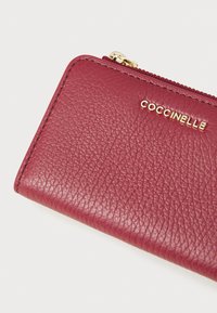 Portafoglio in pelle bordeaux con una finitura texturizzata, chiusura con zip e logo dorato "Coccinelle". Forma rettangolare compatta, interno liscio.