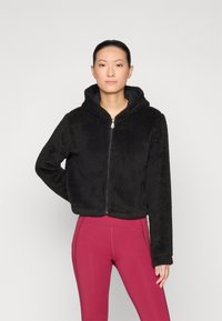 Felpa nera corta realizzata in tessuto morbido e testurizzato, con chiusura a zip e cappuccio integrato, abbinata a leggings rosa aderenti.