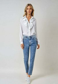 Blouse blanche à volants avec des boutons, associée à un jean taille haute bleu clair et des baskets blanches, le tout sur fond neutre.