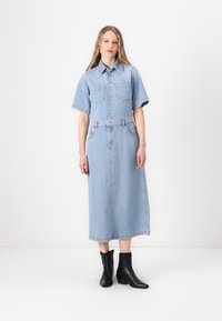 Denim šaty v světle modré barvě. Krátké rukávy, zapínání na knoflíky, dvě náprsní kapsy, pásek v pase a dlouhá, rovná sukně. Černé kotníkové boty.