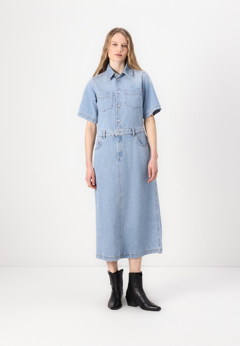 Denim šaty v světle modré barvě. Krátké rukávy, zapínání na knoflíky, dvě náprsní kapsy, pásek v pase a dlouhá, rovná sukně. Černé kotníkové boty.