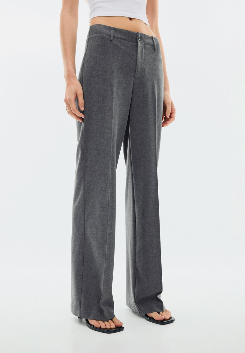 Femme portant un pantalon gris taille haute à jambes larges avec pli devant et sandales noires ouvertes aux orteils, haut court blanc visible au-dessus de la taille.