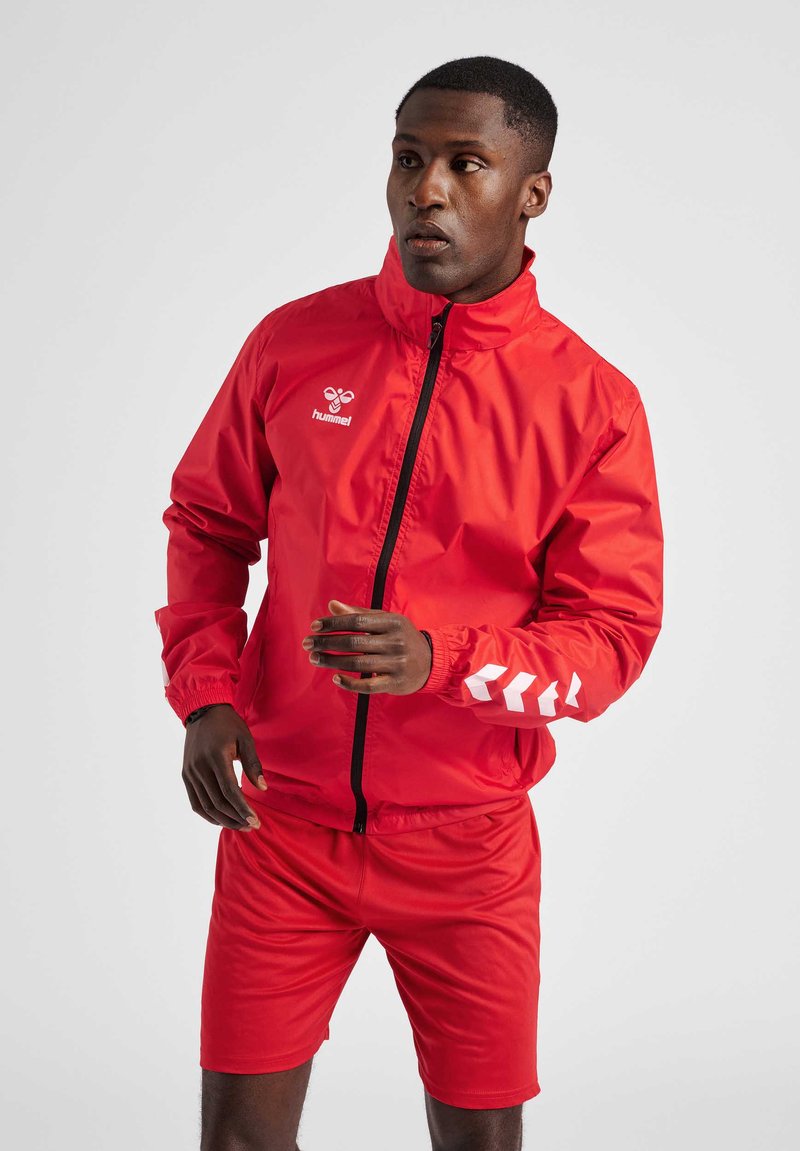Rote Windjacke mit hohem Kragen und Reißverschluss, ausgestattet mit weißem Logo und Chevron-Muster an den Ärmeln. Kombiniert mit roten Shorts.