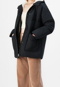 Doudoune noire avec une capuche doublée en fausse peau lainée, dotée d'une fermeture éclair frontale, d'une fermeture à boutons et de grandes poches latérales. Associée à un pantalon en velours côtelé.