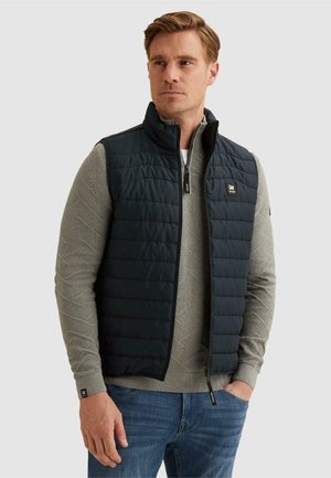 Homme portant une veste matelassée noire sur un pull zippé gris texturé et un jean bleu, debout, une main dans la poche.