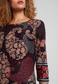 Dlouhý rukáv top v tmavě námořnické modři s propracovanými květinovými a paisley vzory v červené, krémové a modré barvě, s kulatým výstřihem a strukturovanou tkaninou.