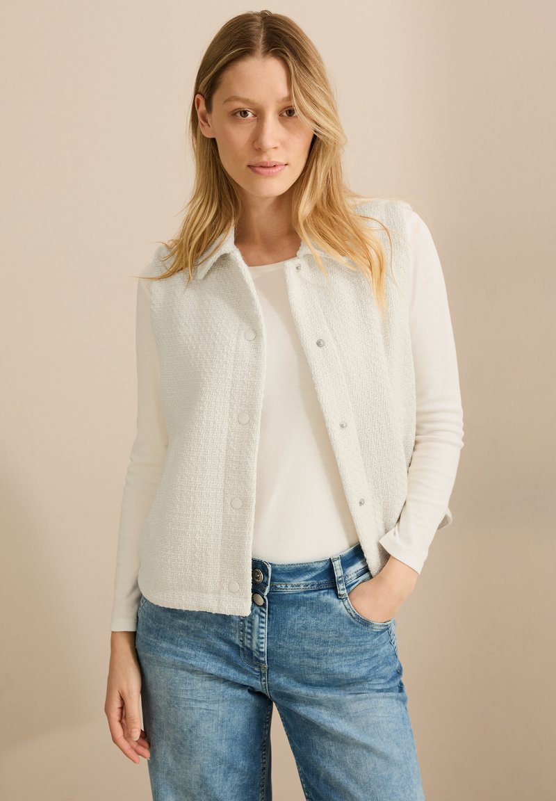 Ljusgrå, texturerad väst med krage och tryckknappar, bärs över en vit långärmad topp och blå jeans.