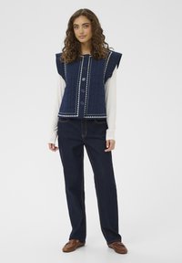 Gilet trapuntato blu navy con spalle larghe, chiusura con bottoni e cuciture decorative bianche, abbinato a pantaloni di denim scuro e mocassini marroni.