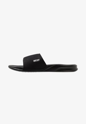 Black slide sandal made of soft fabric with a perforated upper, featuring a thick strap with "REEF" logo. Flat rubber outsole.