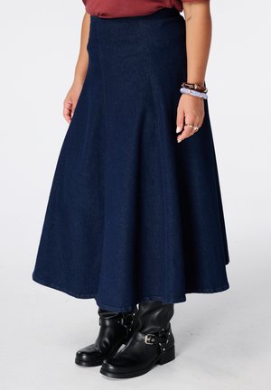 Vrouw draagt een donkerblauwe A-lijn spijkerrok met zwarte enkellaarsjes en een rood topje, toont armbanden en ringen aan haar rechterhand.