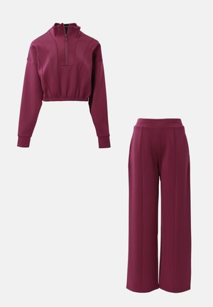 Bordeaux atletiekset met een cropped, rits-top met lange mouwen en hooglopende, wijde broek, beide in een gladde stof.