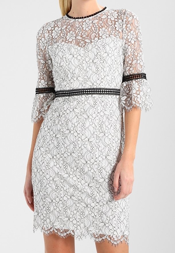 Robe en dentelle blanche à motifs floraux avec une bordure noire au niveau de l'encolure, de la taille et des manches aux coudes, portée par une personne aux cheveux blonds et lisses.