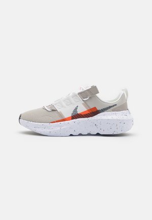Beiger und weißer Sneaker mit gesprenkelter dicker Sohle, orange-schwarzem Nike-Logo und Klettverschluss auf weißem Hintergrund.