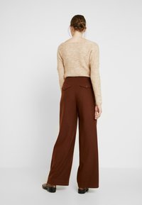 Vrouw met een knot in haar haar, gekleed in een beige gebreide trui, hoog-waist brede bruine broek met knoopzakken aan de achterkant, en slangenleren loafers.