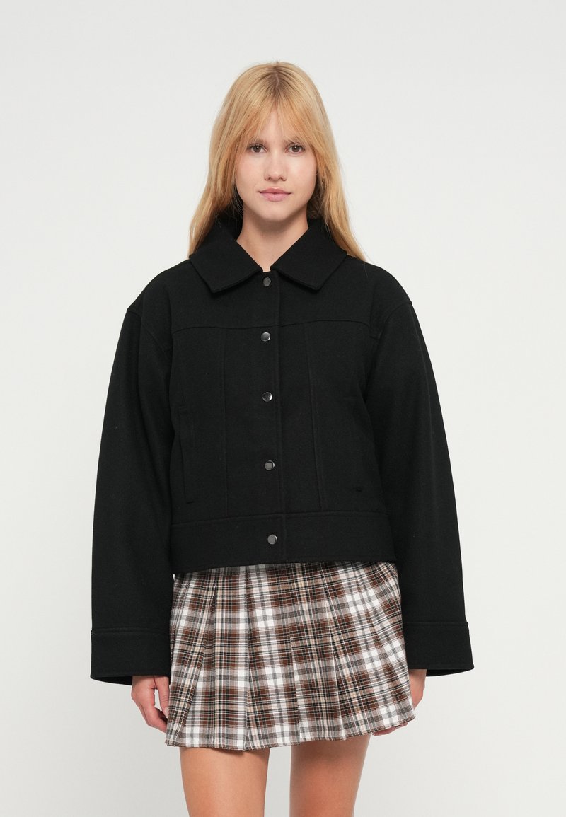 Μαύρο κοντό σακάκι με μυτερό γιακά, κουμπιά μπροστά και πλαϊνές τσέπες, συνδυασμένο με καρό πλισέ φούστα σε καφέ και λευκές αποχρώσεις.