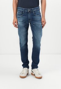 Blå denimjeans med en tapered fit, der har falmende mønstre, fem lommer og en frontknaplukning. Sammen med hvide sneakers.