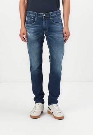 Blå denimjeans med en tapered passform, med blekningsmönster, fem fickor och en framknappsstängning. Matchas med vita sneakers.