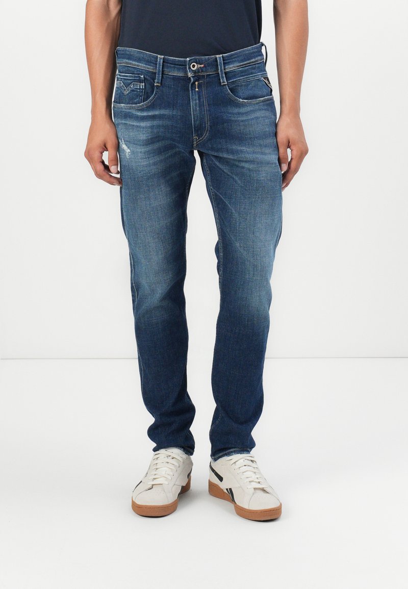 Blå denimjeans med en tapered fit, der har falmende mønstre, fem lommer og en frontknaplukning. Sammen med hvide sneakers.
