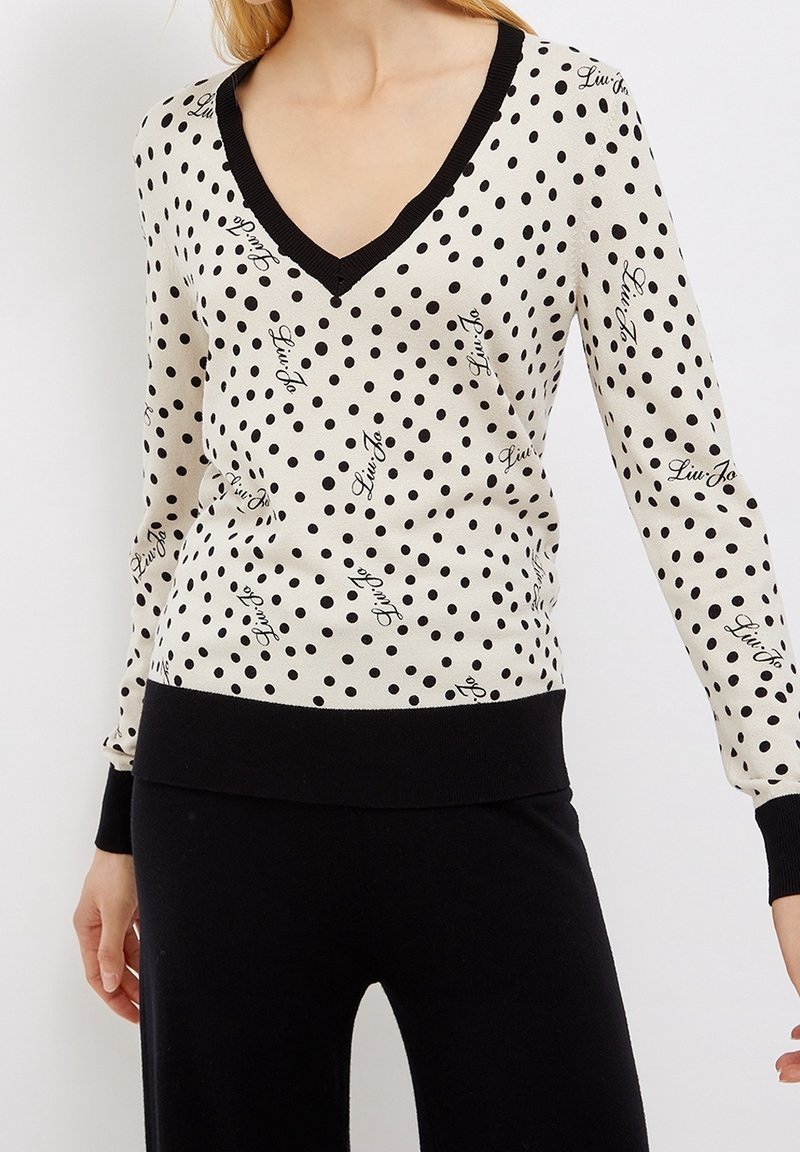 Femme portant un pull blanc à col en V avec des pois noirs et l'inscription « Liu Jo », assorti à un pantalon noir.