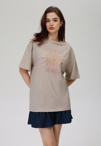 Beige oversized t-skjorte med et solmotiv i rosa og gult. Korte ermer, rund hals, glatt stoff. Kombinert med et denimskjørt.