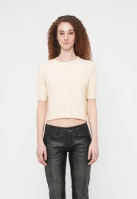 Γυναίκα με σγουρά κόκκινα μαλλιά που φοράει μπεζ κοντομάνικο φερμουάρ κορμάκι και μαύρο υφασμάτινο παντελόνι, στέκεται μπροστά σε λευκό φόντο.