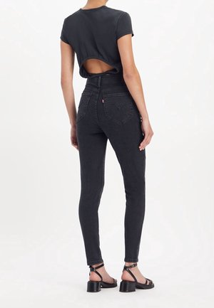 Femme portant un jean skinny taille haute noir avec des poches arrière en forme de cœur, un top noir à manches courtes avec un dos ouvert et des sandales noires à talons.