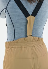 Tan bretelpantalon met elastische tailleband en verstelbare bandjes, gecombineerd met een blauwe textuurtop met donkere stikseldetails.