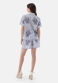 Completo composto da una camicia bianca a maniche corte e pantaloncini, caratterizzato da motivi floreali e a pois blu navy, con bordi scallop. Indossato con sandali con zeppa.