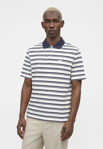 Άνδρας με κοντά ξανθά μαλλιά φοράει λευκό πόλο με μπλε οριζόντιες ρίγες και μπεζ παντελόνι, στέκεται μπροστά σε απλό φωτεινό φόντο.