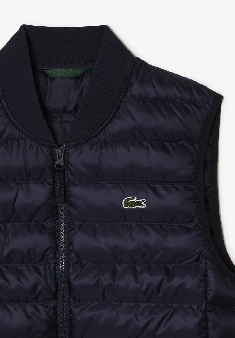 Gilet rembourré bleu marine avec col côtelé, doté d'une fermeture éclair frontale et d'un petit logo brodé. Texture lisse avec un design matelassé.