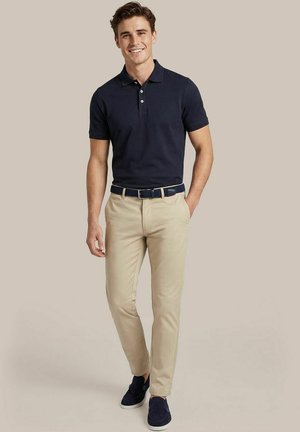 Lachende man staand, dragend een marineblauw poloshirt, beige broek met riem en marineblauwe instapschoenen tegen een effen achtergrond.