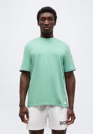 Άνδρας που στέκεται με πρόσωπο προς τα εμπρός, φοράει μια βεραμάν πράσινη μπλούζα και λευκά σορτς με το κείμενο "BOSS" στο αριστερό πόδι, πάνω σε λευκό φόντο.