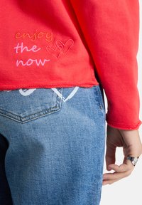 Roter Sweatshirt mit gesticktem Text "Genieß den Moment" und zwei Herzen, kombiniert mit blauer Jeans und einer Hand, die einen silbernen Ring trägt.