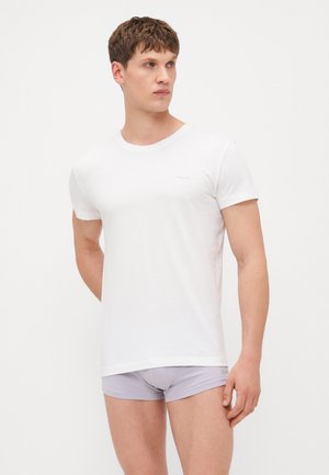 T-shirt blanc à manches courtes en matériau doux, avec un col rond et un petit logo sur la poitrine. Porté avec un short gris.