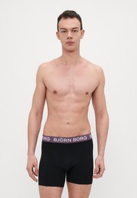 Schwarze Boxershorts mit einem lila Bund, auf dem die Marke "BJÖRN BORG" steht. Der Stoff wirkt glatt mit minimalen Nähten.