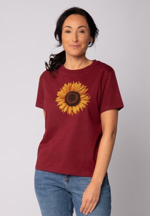 T-shirt en coton bordeaux avec un grand motif de tournesol jaune et marron au centre. Manches courtes et col rond.