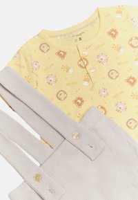 Top de algodón amarillo con estampado de animales, que cuenta con dos botones. Overoles grises de canalé adyacentes con tirantes abotonados y tejido texturizado.