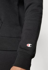 Svart hettegenser med stram mansjett. Har en liten rød og hvit Champion-logo på venstre erme. Myk, teksturert stoff.