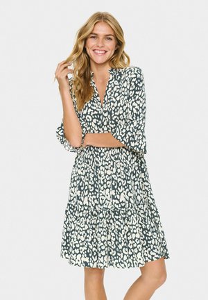 Smiling woman with long blonde hair wearing a black and white animal print knee-length dress, posing with one hand touching hair.